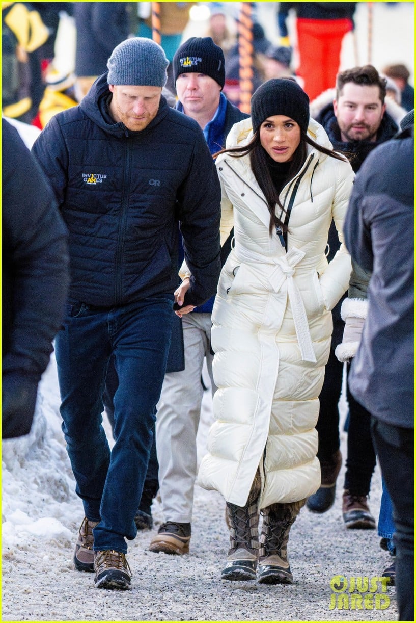 Meghan Markle walking through snow in a long white padded coat beside Prince Harry